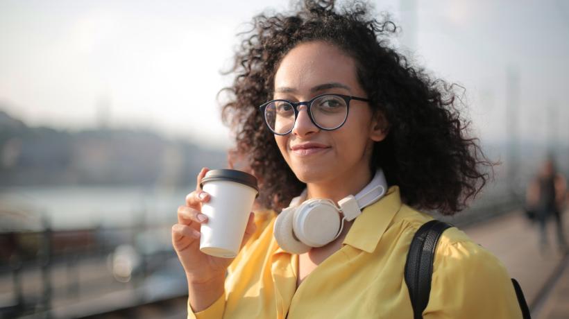 Eine junge Frau hält einen Coffee to go in der Hand, hat die Kopfhörer um ihren Hals gelegt und blickt mit wachen Augen in die Kamera.