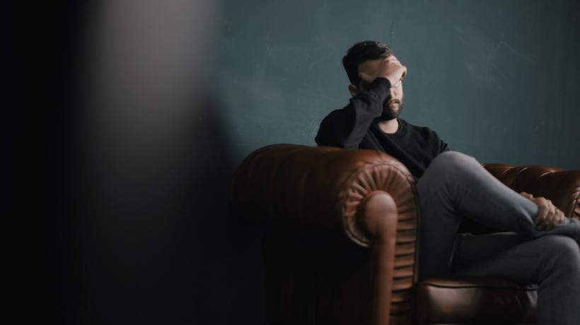 Ein Mann sitzt im Dunklen auf einer Couch und hält eine Hand vors Gesicht.