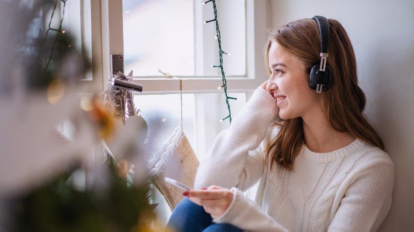 Eine junge Frau sitzt an einem Fenster und hört einen Podcast