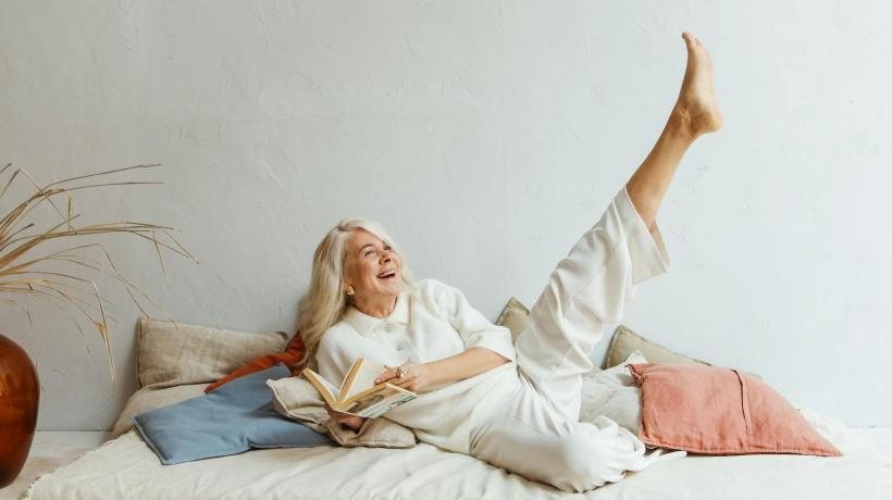 Eine ältere Frau liegt lächelnd auf einem Sofa und streckt ein Bein in die Höhe.