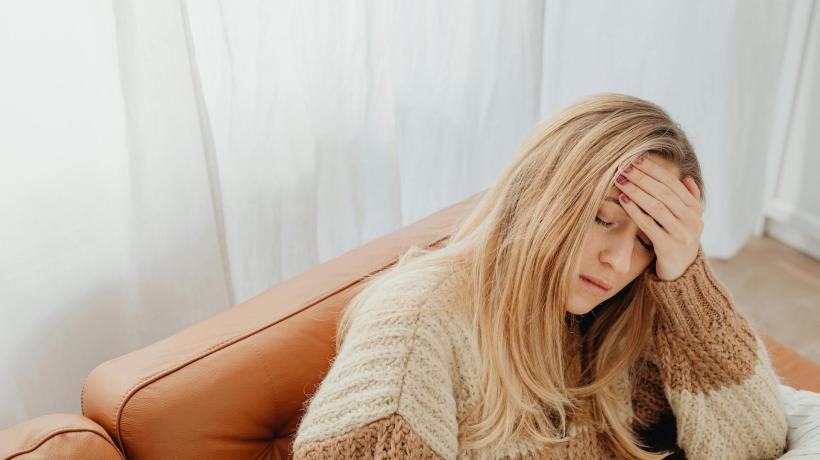 Eine blonde junge Frau sitzt auf einem Sofa und hält sich ihren schmerzenden Kopf.