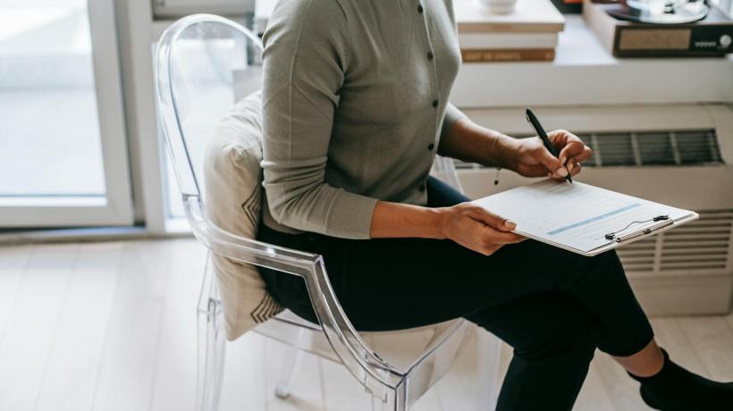 Foto: Alex Green - pexels.com Eine Frau sitzt auf einem Stuhl und füllt mit einem Stift einen Fragebogen aus.