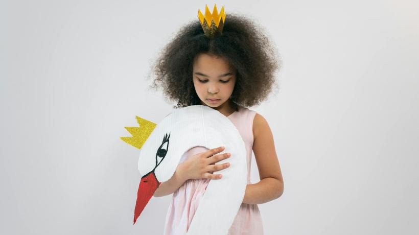 Ein kleines Mädchen hält einen Schwanenkopf aus Papier mit Krone in der Hand und hat selbst eine kleine Krone auf dem Kopf.