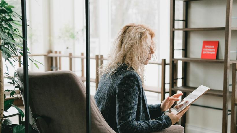 Eine blonde Frau sitzt mit einem Tablet in der Hand in einem Sessel, im Hintergrund ein Regal und Geländer.