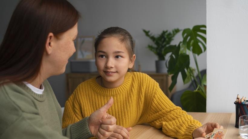 Foto: Freepik.com Ein junges Mädchen sitzt an einem Tisch und schaut zu der neben ihr sitzenden Therapeutin.