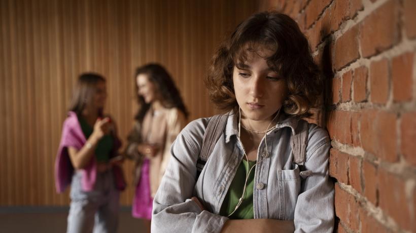 Foto: Freepik.com Ein trauriges Mädchen lehnt an einer Wand, im Hintergrund sieht man zwei sie mobbende Mädchen