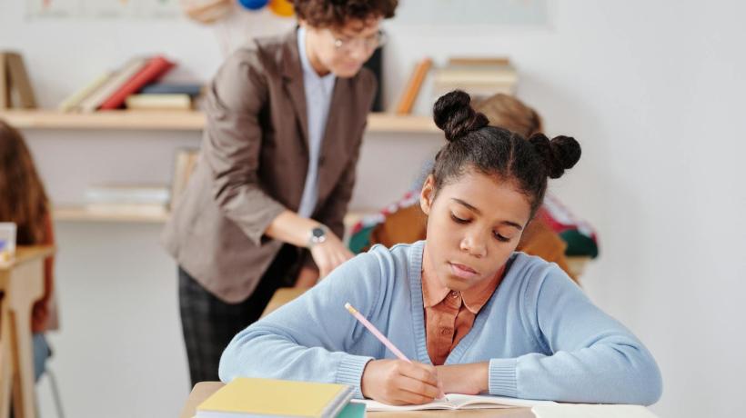 Eine Schülerin sitzt an ihrem Schulpult und bearbeitet eine Aufgabe, ein Lehrer hilft einer Schülerin hinter ihr.