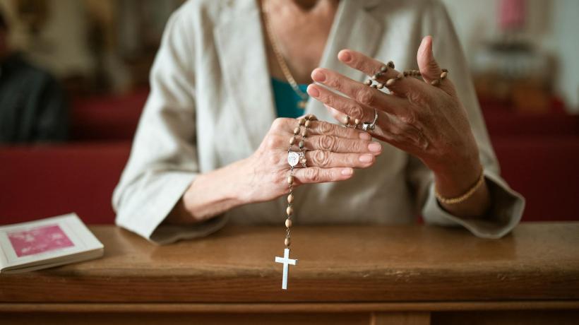 Man sieht eine Frau in Nahaufnahme, die mit einer Perlenkette, an der ein Kreuz hängt, den Rosenkranz betet.