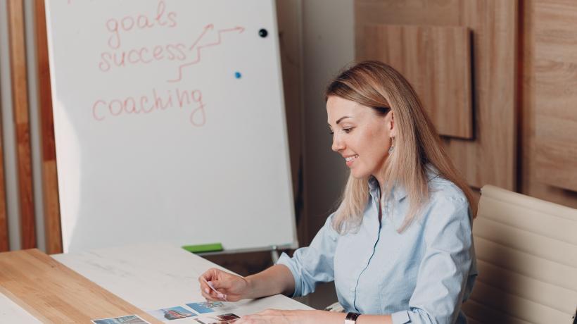 Foto: primipil – stock.adobe.com Eine junge Frau sitzt mit Coaching-Karten an einem Tisch, im Hintergrund ein Whiteboard.