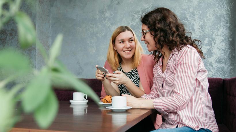 Zwei Frauen sitzen mit je einer Kaffeetasse an einem Tisch und unterhalten sich.