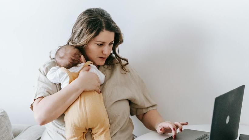 Eine junge Mutter hat ihr Baby im Arm und arbeitet parallel am Laptop