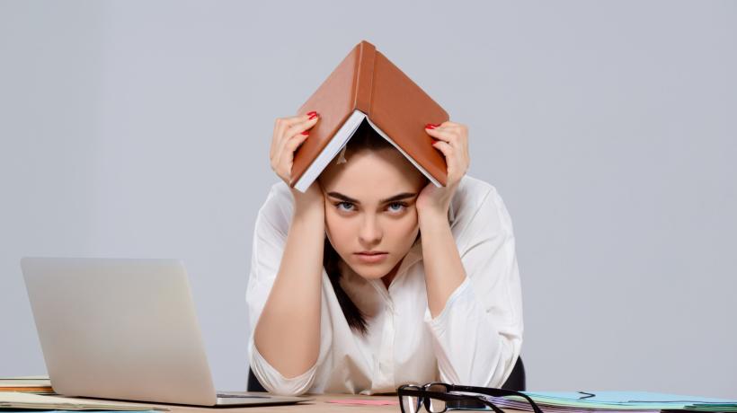 Foto: cookie_studio – freepik.com Eine Frau hält sich frustriert ein Buch über den Kopf und sitzt am Tisch, auf dem ihr Laptop steht.