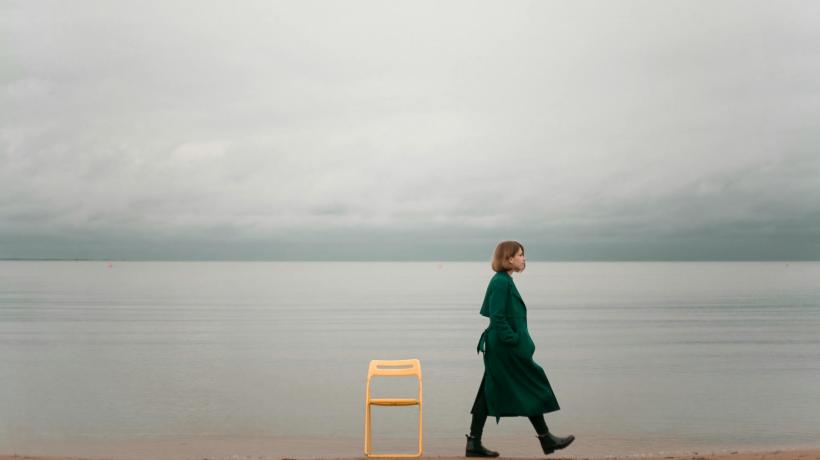 Foto: Polina Sirotina – pexels.com Eine Frau läuft einen leeren Strand entlang, vor dem Meer steht ein gelber leerer Stuhl
