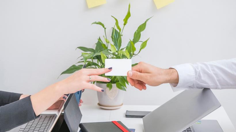 Foto: freepik.com Man sieht die Hände zweier Menschen, die sich an einem Schreibtisch mit Laptops sitzend eine Visitenkarte überreichen.