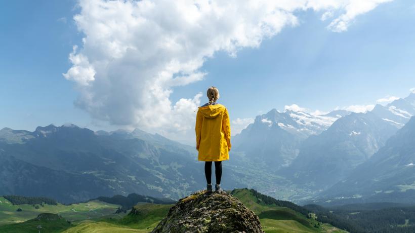 Rückansicht einer Frau in gelber Jacke, die von einer Anhöhe in eine bergige Landschaft schaut