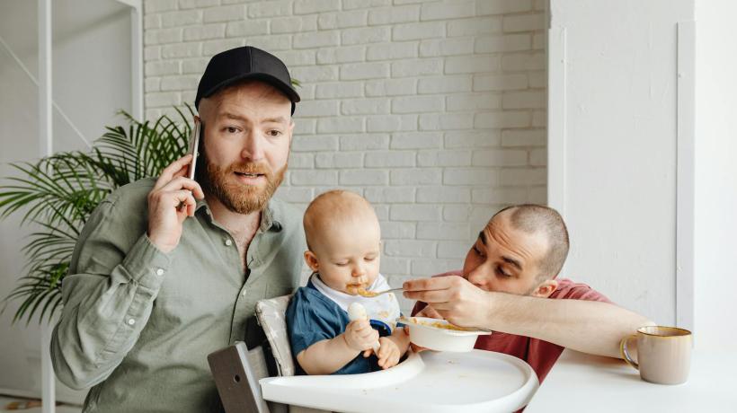 Foto: Kaboompics.com – pexels.com Zwei junge Männer sitzen bei einem Kind im Hochstuhl, der eine füttert das Kind, der andere telefoniert.