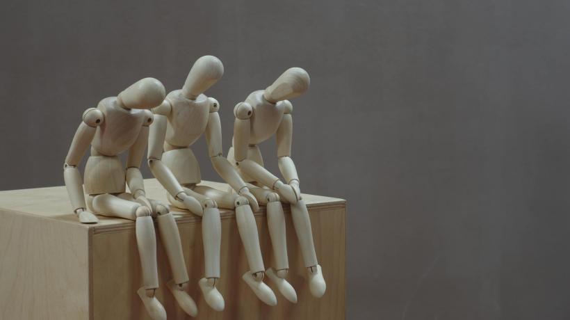 Foto: Marco Bianchetti- pexels.com Drei Holzpuppen sitzen in gebückter, niedergeschlagener Haltung nebeneinander auf einem Klotz.