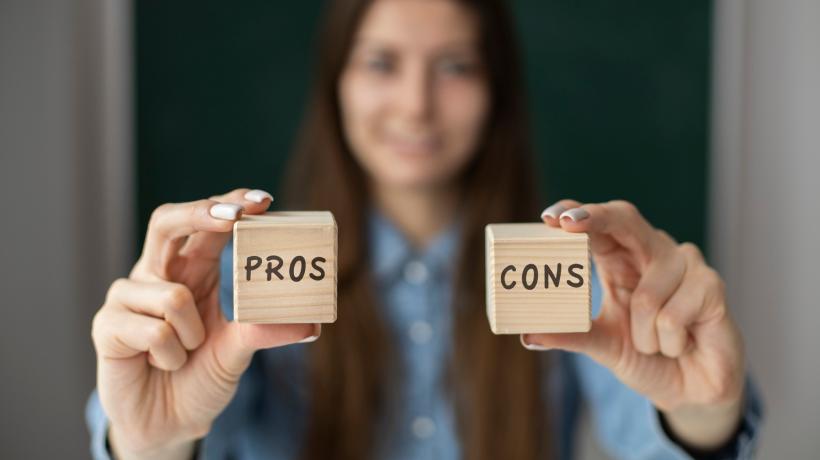 Foto: freepik.com Eine Frau hält zwei kleine Holzwürfel hoch, auf denen einmal PROS und einmal CONS steht.