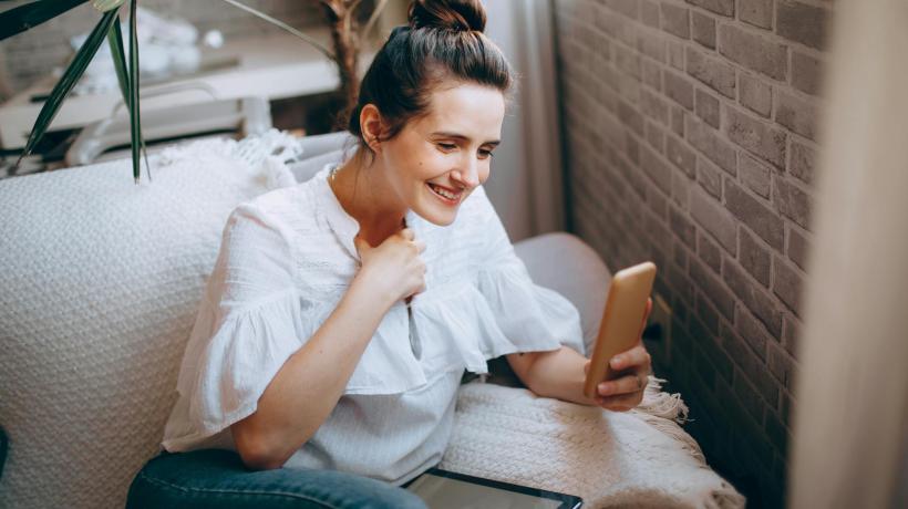 Foto: Helena Lopes – pexels.com Eine junge Frau sitzt auf einer Coach und schaut lächelnd auf ihr Smartphone.