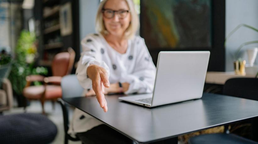 Foto: Yan Krukau – pexels.com Eine Frau sitzt am Laptop und streckt grüßend die Hand Richtung Kamera aus.