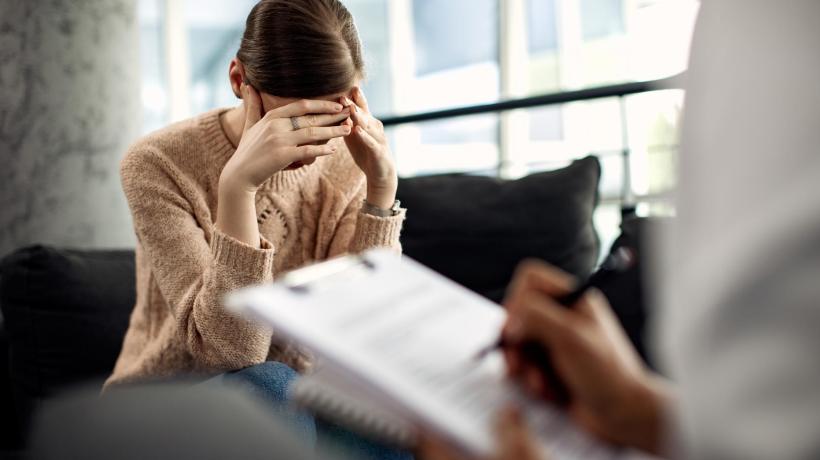 Foto: Drazen Zigic – freepik.com Eine Frau verbirgt ihr Gesicht in den Händen, ihr gegenüber füllt ein:e Psychotherapeut:in einen Fragebogen aus.