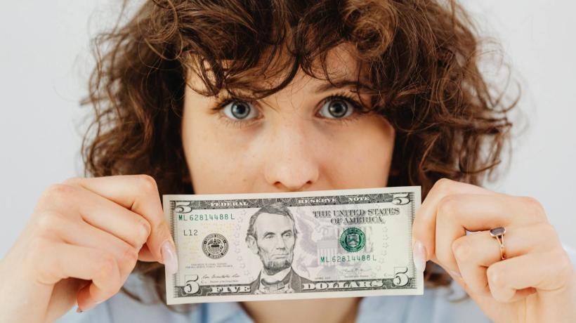 Foto: Kaboompics.com – pexels.com Eine brünette Frau hält sich einen Dollarschein vor das Gesicht.