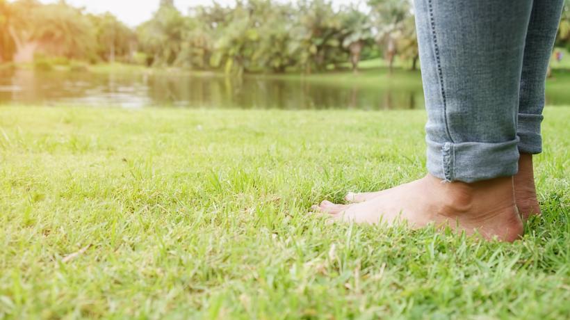 Foto: mindandi – freepik.com Man sieht zwei nackte Füße einer Person, die auf einer grünen Wiese steht, im Hintergrund ein See und Bäume.