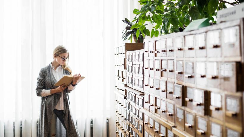 Foto: freepik.com Eine Frau steht eine Akte lesend vor einem großen Aktenschrank.