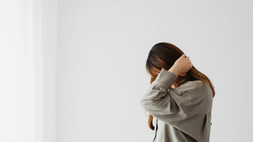 Foto: Kaboompics.com – pexels.com Eine Frau verbirgt das Gesicht in den Händen und steht vor hellem Hintergrund.