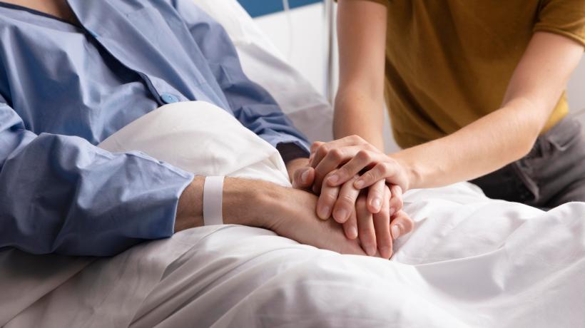 Eine junge Person hält ihre Hände auf die verschränkten Hände einer alten Person, die in einem Bett liegt.