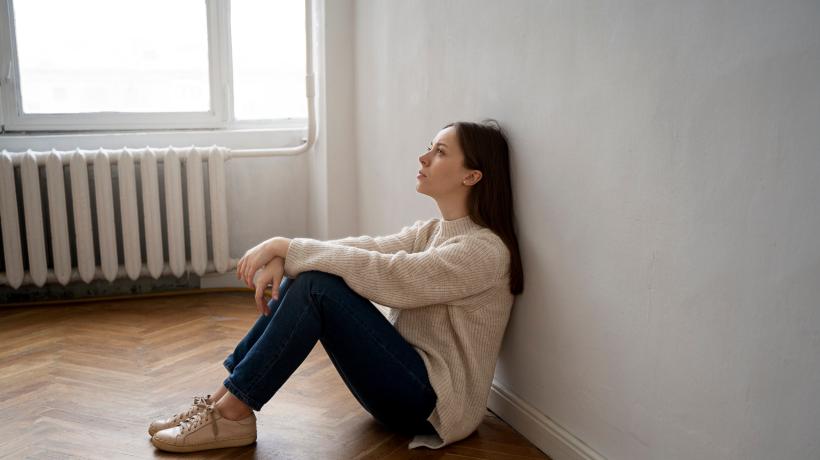 Eine dunkelhaarige junge Frau sitzt mit dem Rücken an einer Wand auf dem Boden und schaut müde.