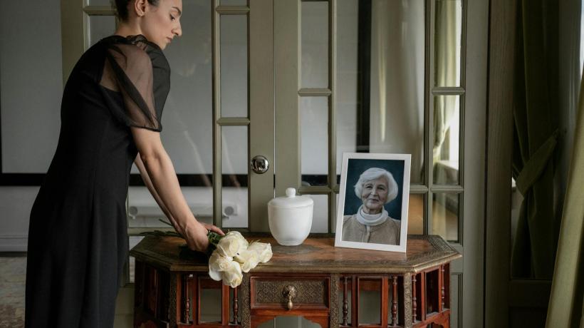 Eine Frau in Trauerkleidung steht an einem Gedenktisch, auf dem ein Bild, weiße Blumen und eine Urne stehen