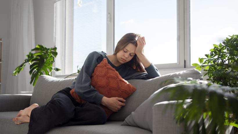 Eine brünette Frau sitzt auf einem Sofa und hält sich müde ein Kissen vor den Bauch.
