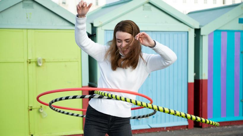 Eine Frau balanciert vor bunten Häusern mit drei Hulahoop-Reifen