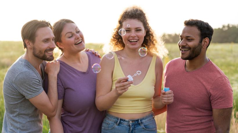 Foto: freepik.com Vier junge Leute stehen lachend und mit Seifenblasen nebeneinander vor einer sommerlichen grünen Landschaft.
