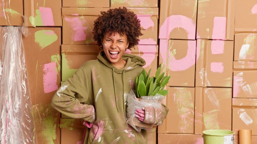 Eine junge Frau mit dunklen Haaren steht vor einer bemalten Wand in farbbekleckster Kleidung und hält lächelnd eine Topfpflanze in der Hand.