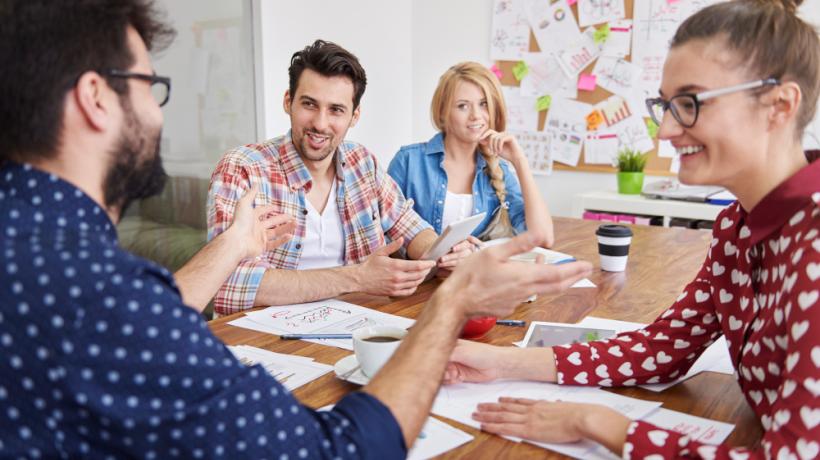 Zwei Frauen und zwei Männer unterhalten sich an einem Bürotisch, im Hintergrund sieht man eine Pinnwand.