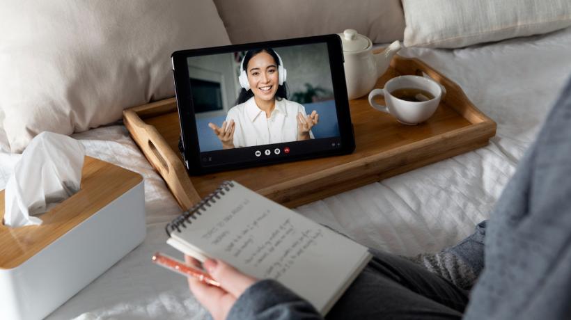 Man sieht die Hände einer Person, die einen Notizblock in der Hand hält und auf dem Bett vor einem Tablet sitzt.