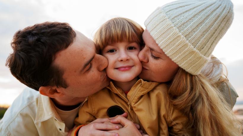 Ein Kind zwischen seinen Eltern, die ihm je einen Kuss auf die Wange geben.