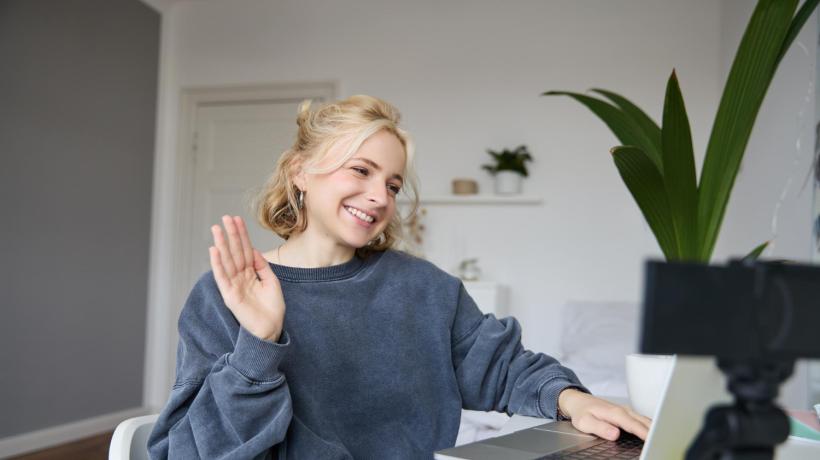 Eine blonde junge Frau sitzt vor dem Laptop und winkt zum Bildschirm.