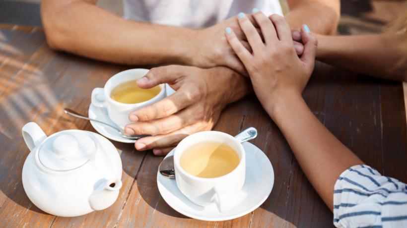 Zwei Menschen sitzen mit zwei Kaffees und übereinandergelegten Händen an einem Holztisch