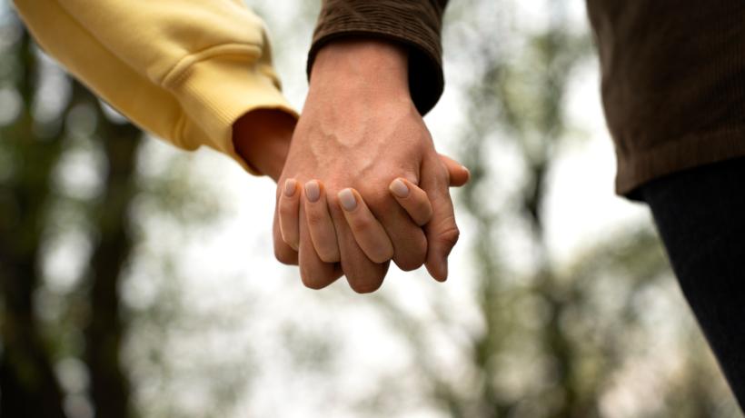Nahaufnahme der einander umschließenden Hände zweier Personen, im Hintergrund sind Bäume zu sehen.