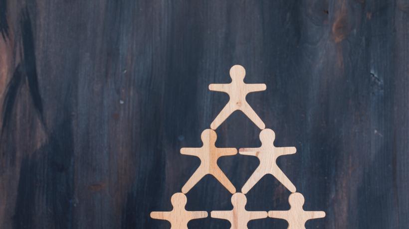 6 zu einer Pyramide aufeinandergestellte Holzmännchen vor dunklem Hintergrund.