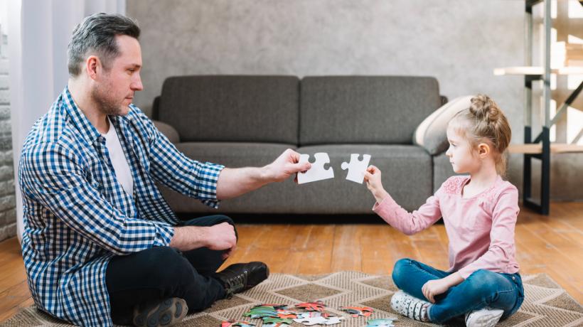 Ein Mann und ein kleines Mädchen sitzen auf dem Boden vor einem Puzzle und führen zwei Puzzleteile in der Luft zusammen.