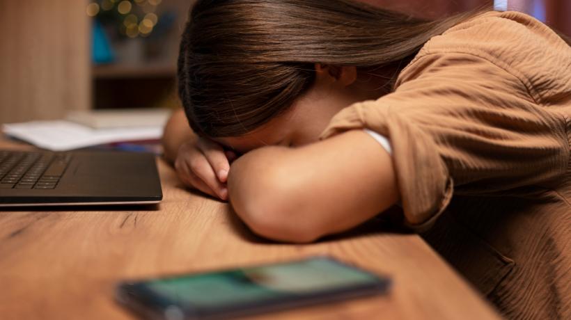 Ein Mädchen sitzt an einem Tisch und hat den Kopf auf die Hände gelegt und versteckt ihr Gesicht. Neben ihr liegt ein Smartphone.