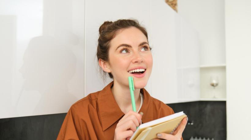 Eine junge Frau sitzt mit Schreibblock auf einem Sofa und überlegt lächelnd.