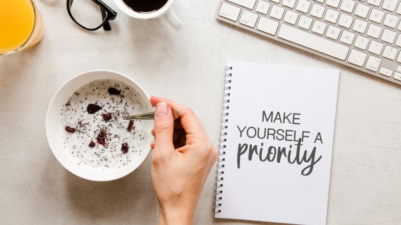 Vogelperspektive auf einen Schreibtisch mit Kaffeetasse, einem Block mit Aufdruck "Make yourself a priority", Brille, Tastatur, Saftglas