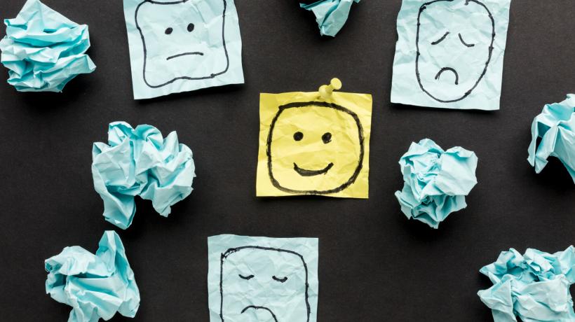 Vier Post-its, drei davon mit negativen oder neutralen Smileys, ein gelber mit lächelndem Smiley. Drumherum zerknüllte Papiere.