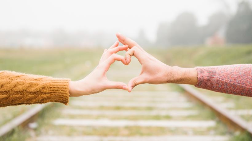 Die Hände von zwei Personen formen ein Herz, auf dem Boden sieht man Bahngleise.