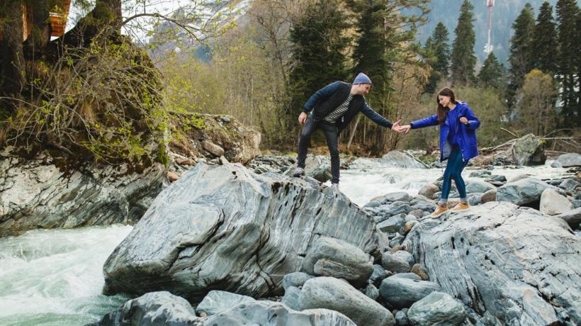 Ein Mann reicht einer Frau helfend die Hand, damit sie auf einen Felsen an einem Fluss klettern kann.